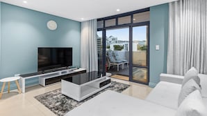 Una sala de estar moderna con un televisor de pantalla plana, un sofá blanco y un puerta de vidrio que da a un balcón con mobiliario exterior.