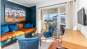 Una sala de estar moderna con un sofá de cuero, un sillón azul, una mesa de comedor de madera y un ventanal que da a un balcón con vistas a edificios.