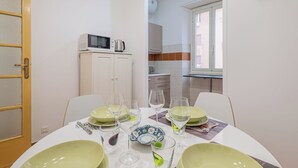 A dining area with a table set for four, including plates, glasses, and cutlery. A microwave is visible in the background, and the room has a simple, clean design with white walls and a window.