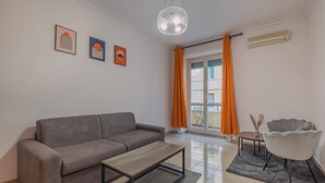 Ein modernes Wohnzimmer mit einem grauen Sofa, einem Holztisch und einem grauen Sessel. An der Wand hängen drei gerahmte Kunstwerke, eine Glühbirne in der Decke und ein Fenster mit orangefarbenen Vorhängen.
