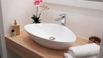 A modern bathroom with a white oval sink, a wooden vanity, a small plant with pink flowers, and a white towel on the counter.