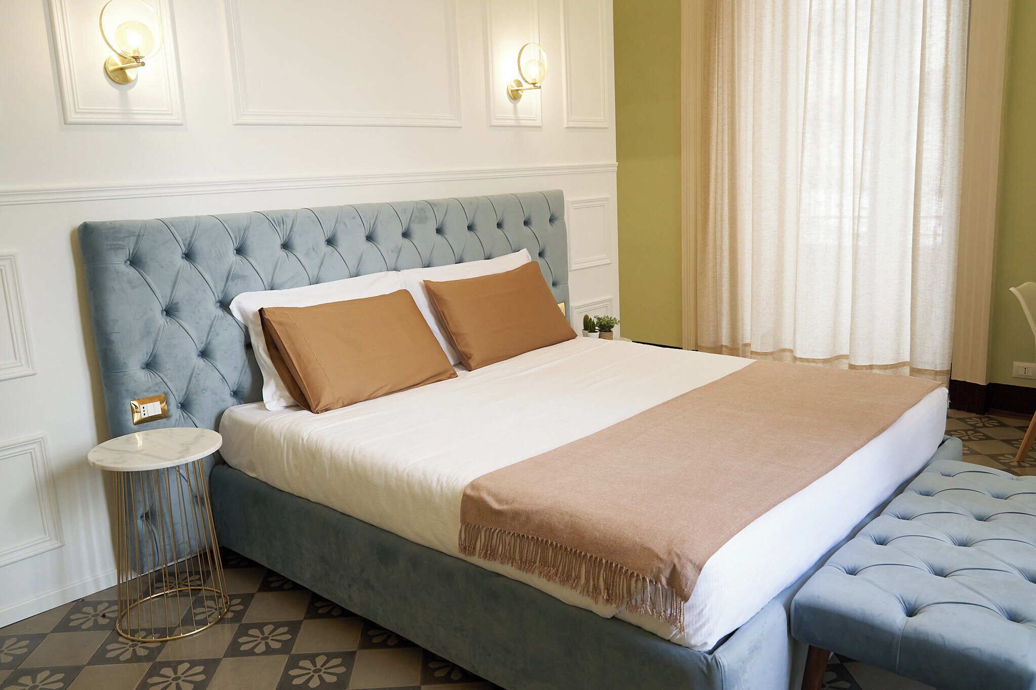 Een slaapkamer met een gestuukt hoofdeinde, een rond bijzettafeltje, een bankje en een bed met een beige en bruine deken.