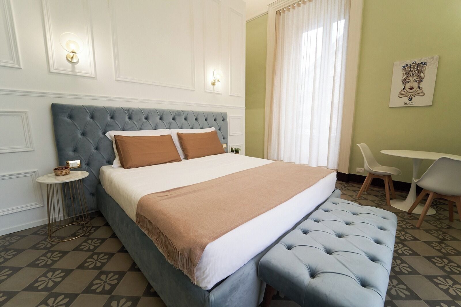 A neatly arranged bedroom with a tufted headboard, a large bed with two pillows, a bedside table with a basket, and a small round table with chairs.