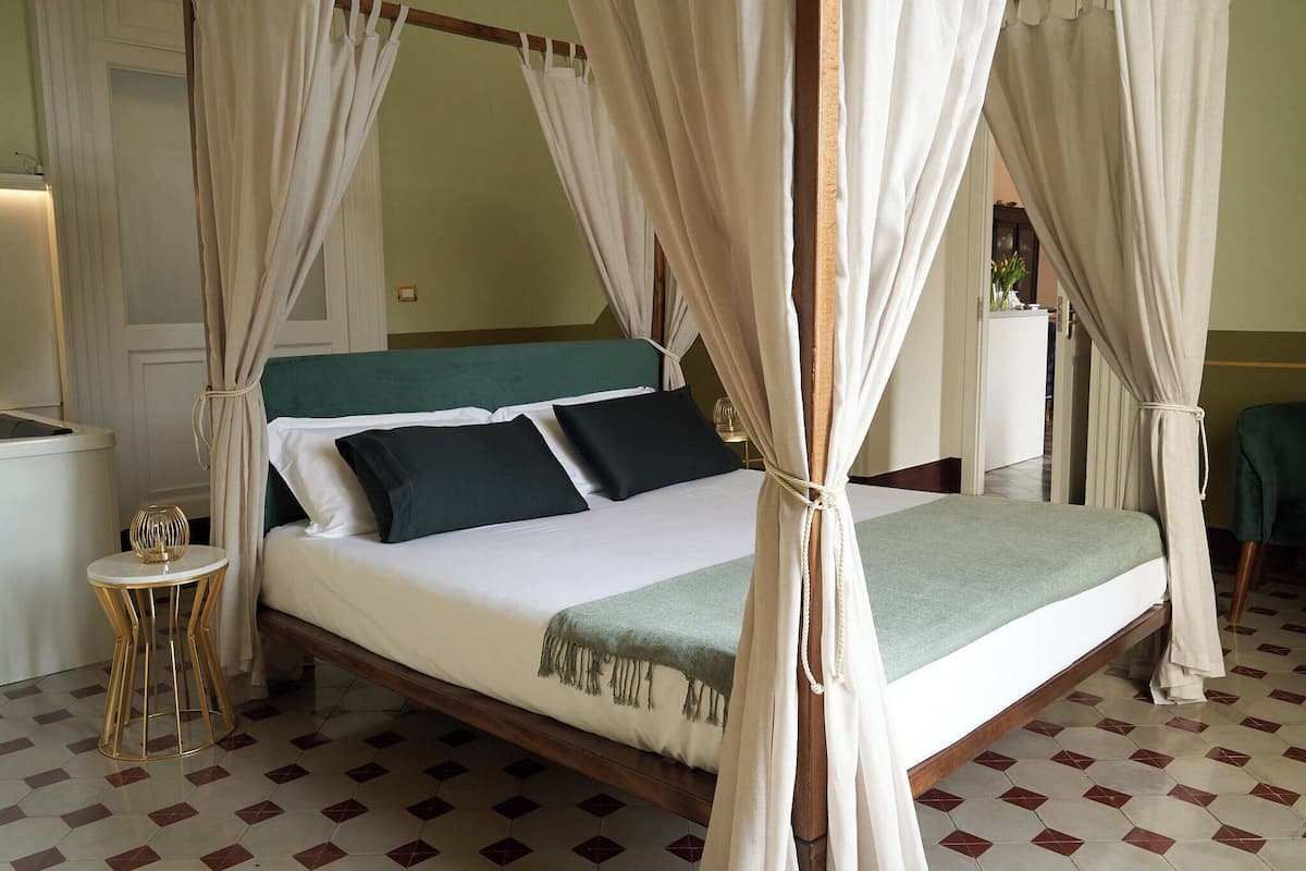 A four-poster bed with a green headboard, white bedding, and a green throw blanket. There is a small round table with a gold base and a decorative item on it. The room has a checkered floor pattern.