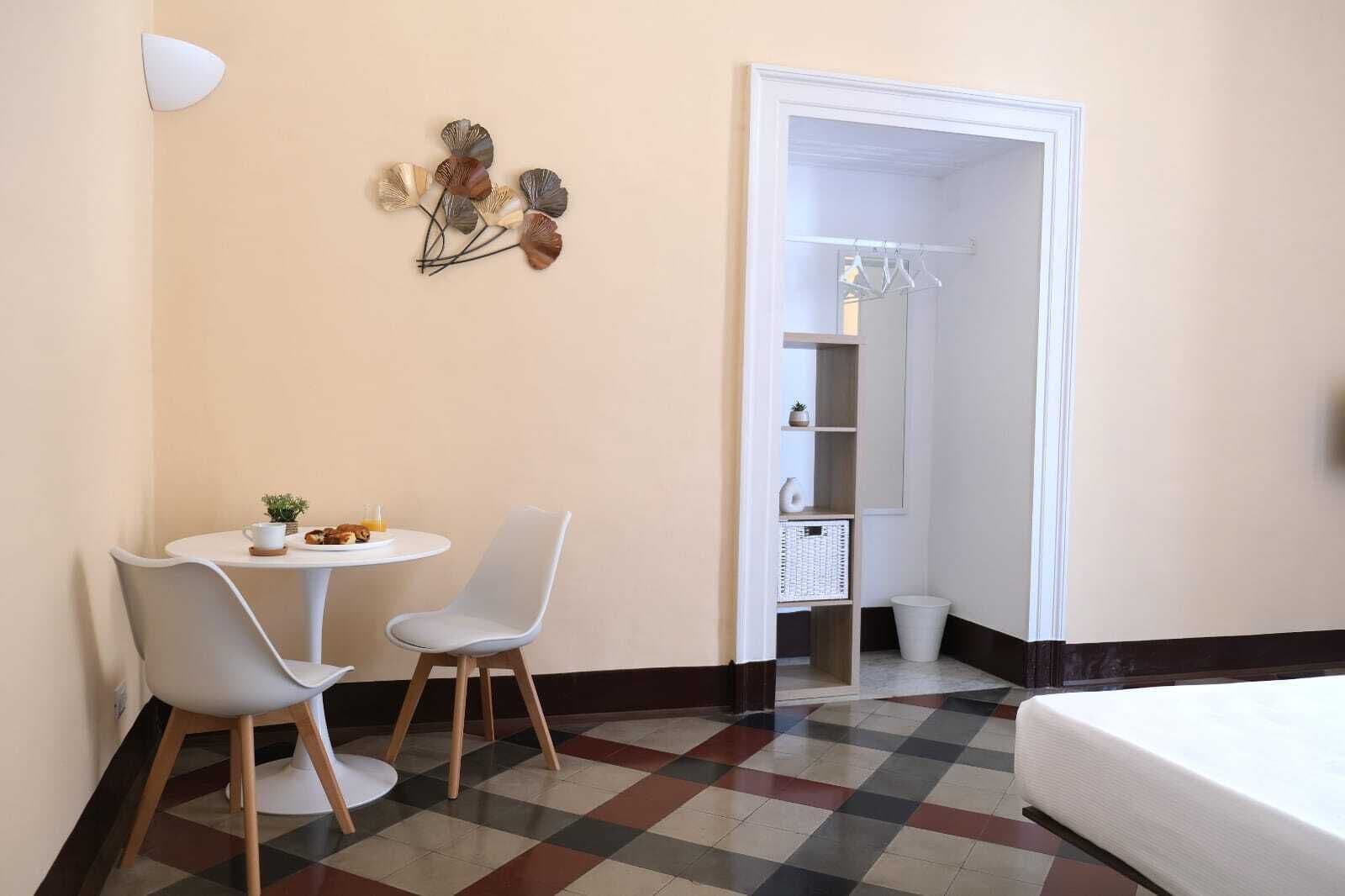 Habitación de hotel con piso a cuadros, una cama, una pequeña mesa con sillas, un aplique de pared y un armario con ropa colgada.