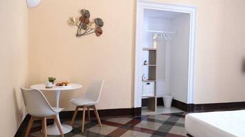 Habitación de hotel con piso a cuadros, una cama, una pequeña mesa con sillas, un aplique de pared y un armario con ropa colgada.