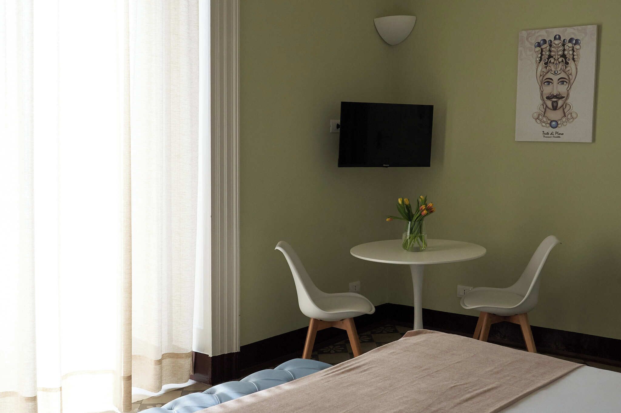 A hotel room with a bed, two white chairs, a small round table with a vase of flowers, a flat-screen TV mounted on the wall, and a framed artwork above the TV.