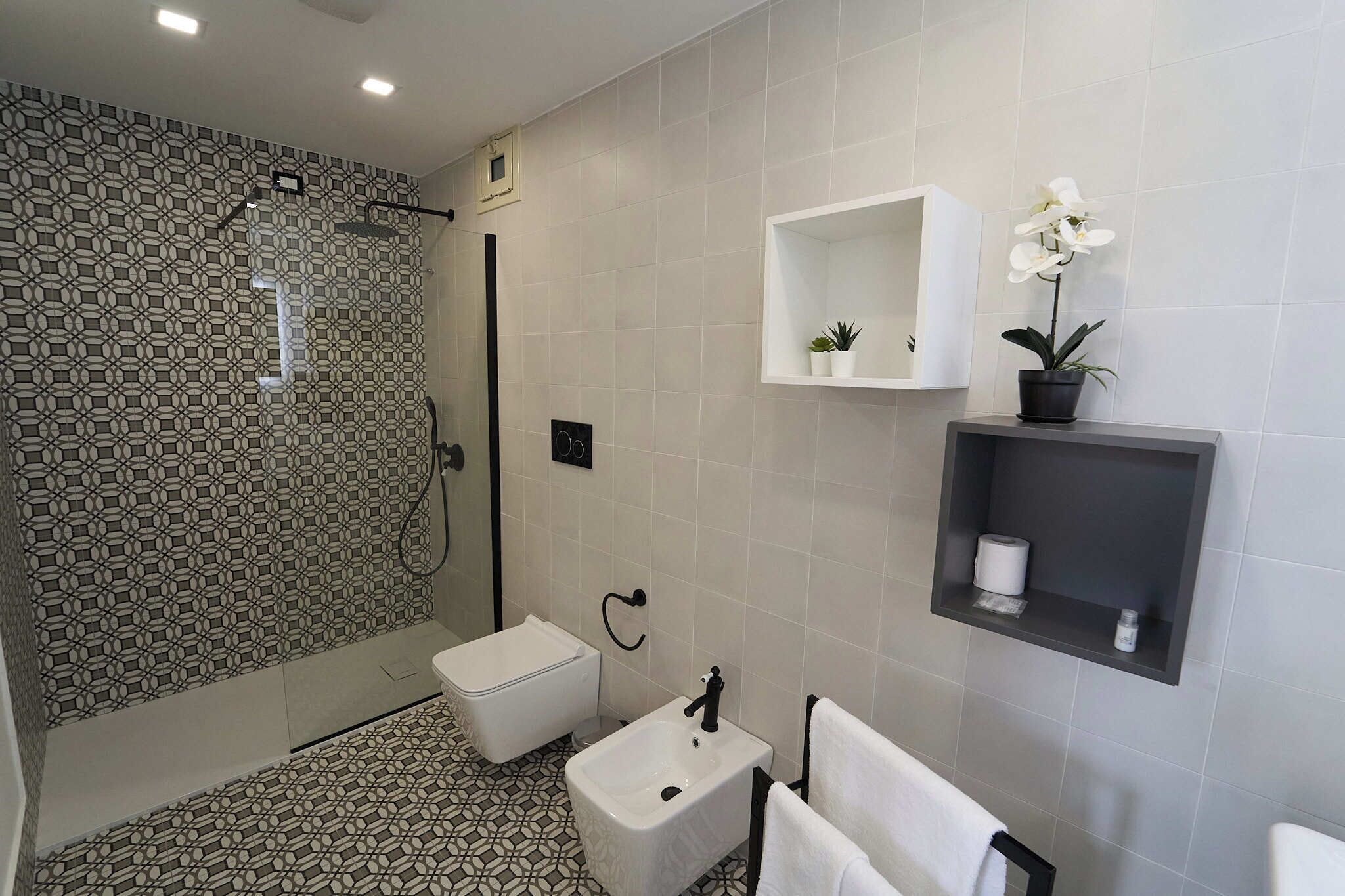 A modern bathroom with a patterned shower wall, a white sink, a toilet, and a shelf with a potted plant.