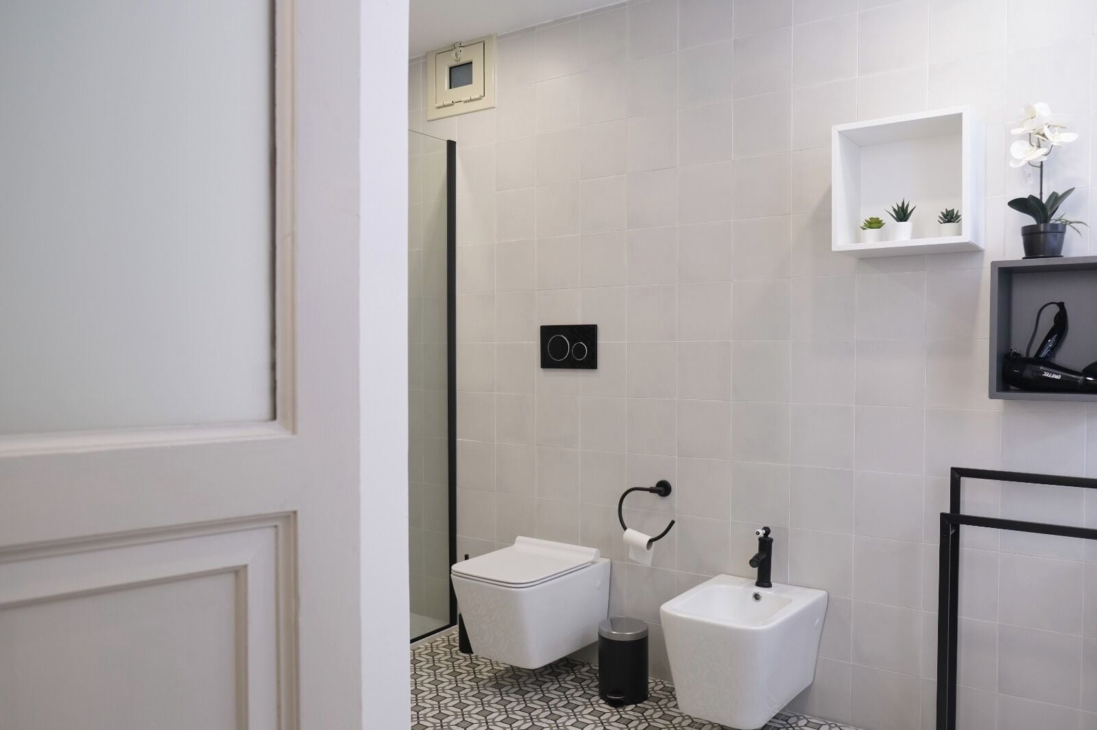A modern bathroom with a toilet, bidet, and sink. There are potted plants on a shelf and a trash bin on the floor.