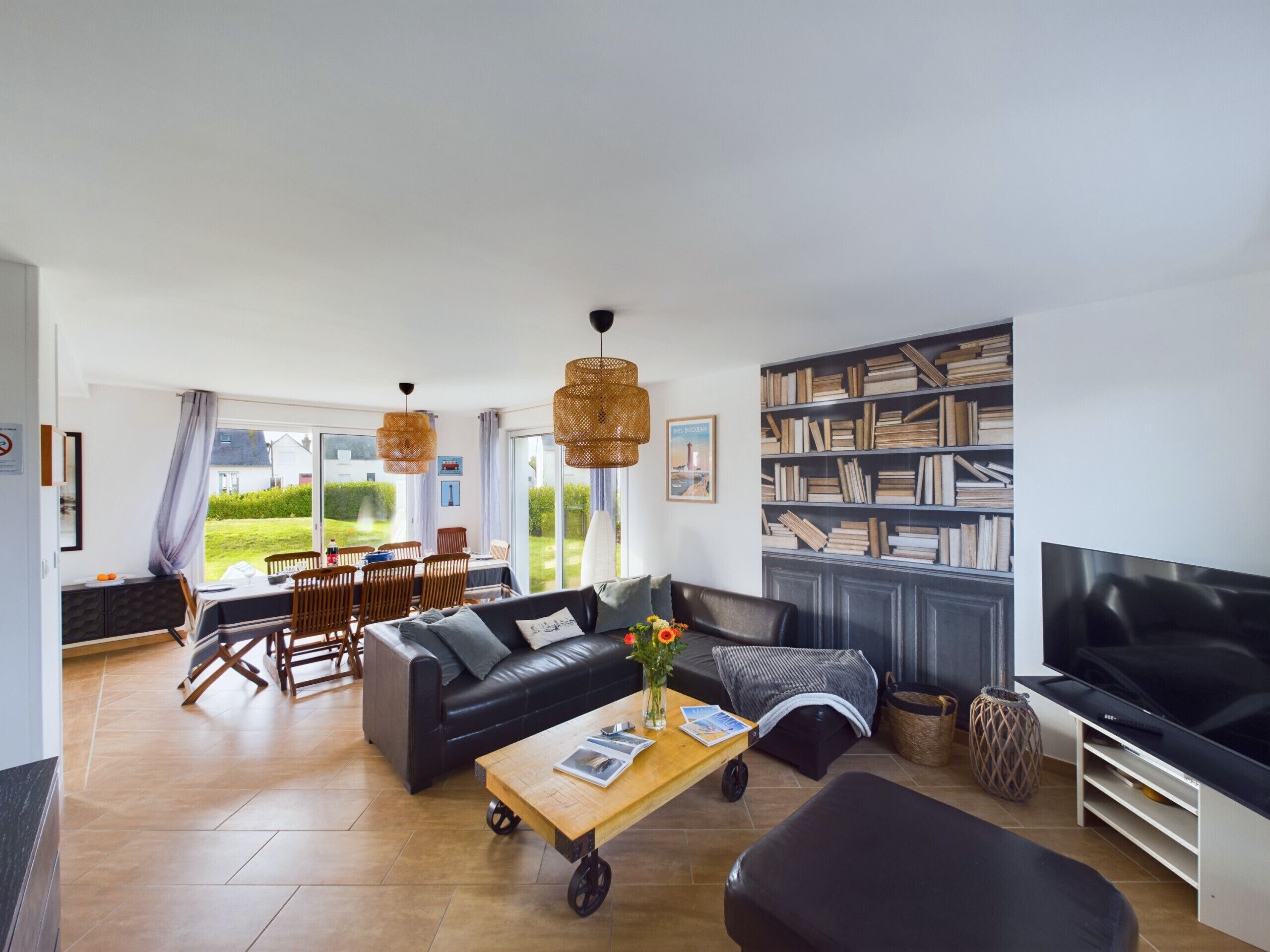 Una sala de estar moderna con un sofá, una mesa de centro y un televisor. Hay una zona de comedor con sillas y una mesa, y una estantería llena de libros.