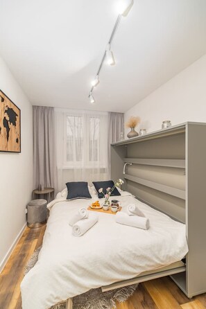 Une chambre à coucher moderne avec un lit, des tables de chevet, une fenêtre avec des rideaux, un petit tabouret et une poubelle.