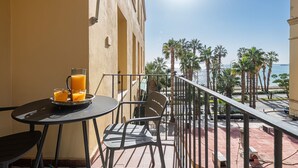 Un balcón con mesa y sillas, un vaso de jugo de naranja, y vistas a palmeras y al mar.