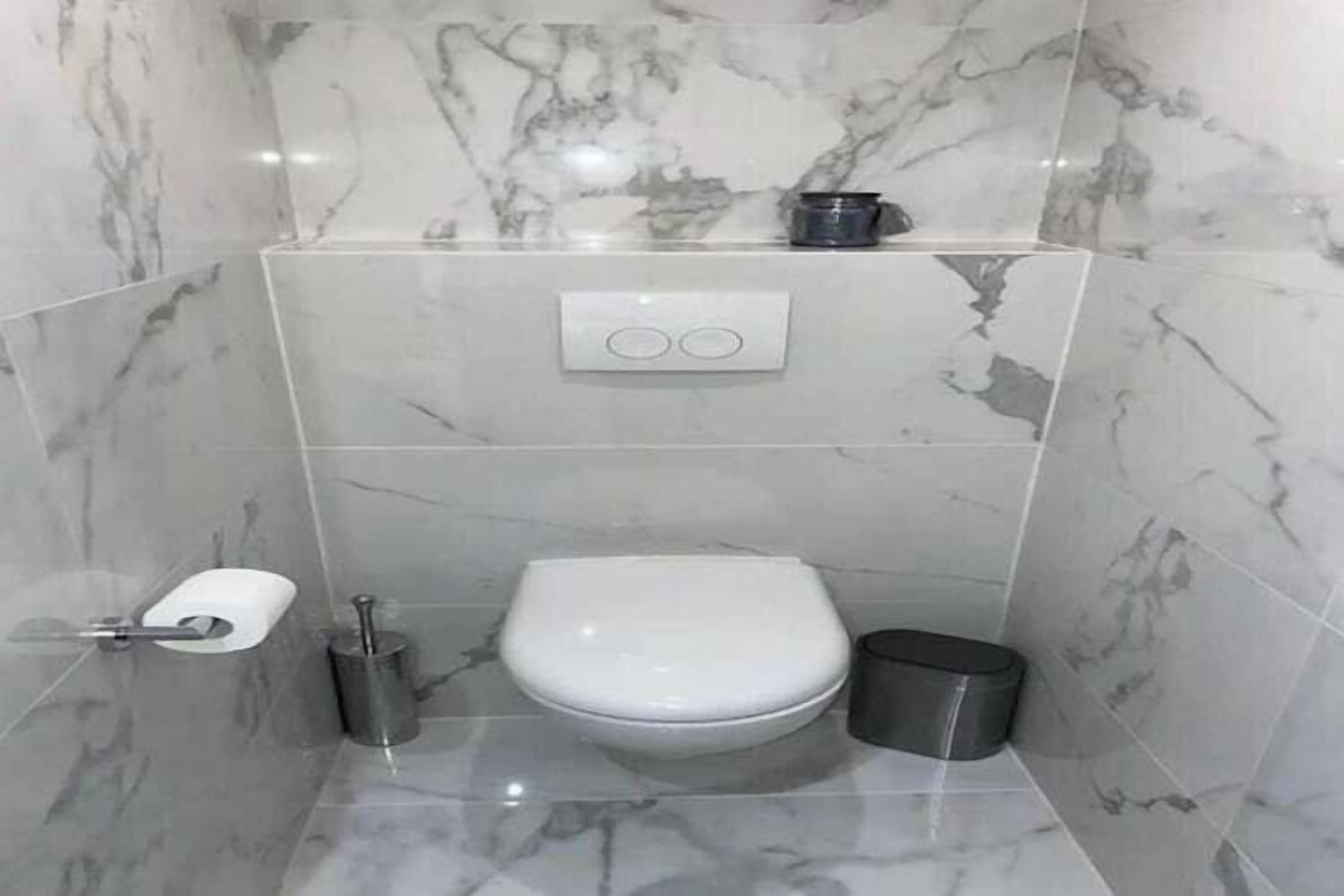 Une salle de bain avec des toilettes blanches, un pare-chocs en marbre, une petite étagère avec une tasse et une poubelle.