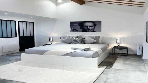 A modern bedroom with a large bed, a ceiling with wooden beams, and a large window with blinds.