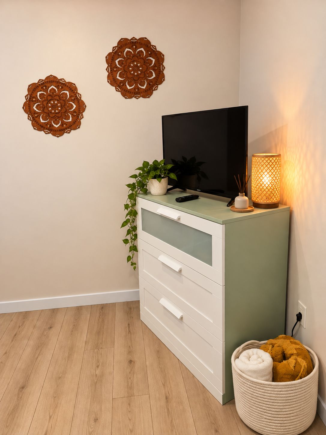 Une pièce avec un téléviseur posé sur un meuble, des œuvres d’art décoratives accrochées au mur, une plante en pot, une lampe et un coffre de rangement contenant des coussins.