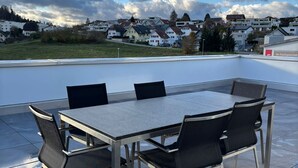 Une terrasse sur le toit, avec une table rectangulaire et six chaises, d’où l’on a une vue sur un quartier résidentiel avec des maisons et des arbres.