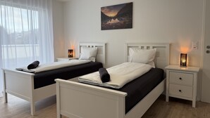 Une chambre d’hôtel avec deux lits, une table de chevet avec une lampe, un tableau au mur et une fenêtre avec des rideaux légers.