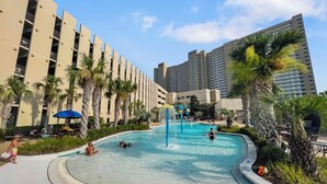 A swimming pool with a water fountain, surrounded by palm trees and lounge chairs, with tall buildings in the background. - Emerald Beach Resort 2028 (Panama City Beach)