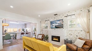 Uma sala moderna com um sofá amarelo, uma poltrona de couro marrom e uma lareira. Há uma área de jantar com mesa e cadeiras, e uma cozinha ao fundo.