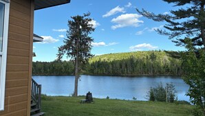 A wooden cabin with a deck overlooking a lake, surrounded by trees and a clear sky.