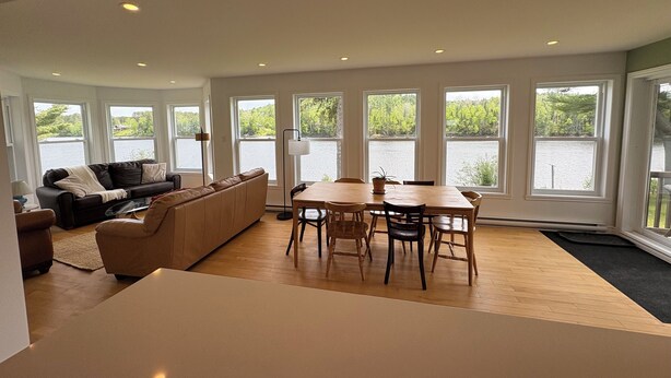 A spacious living area with a large window overlooking a lake, a wooden dining table with chairs, and a beige sofa.