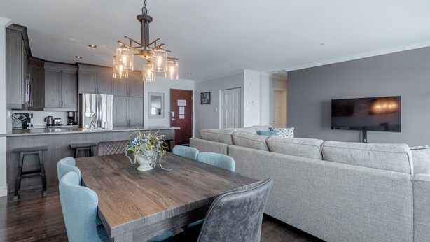 A modern living room with a grey sofa, a wooden dining table with four chairs, a kitchen area with grey cabinets, and a flat-screen TV mounted on the wall.