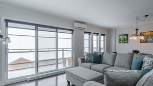 A modern living room with a grey sectional sofa, large windows, and an air conditioning unit.