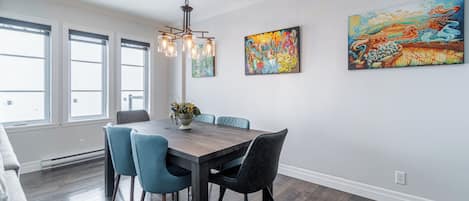 A dining area with a wooden table, blue chairs, and a chandelier. There are two large windows and a painting on the wall.