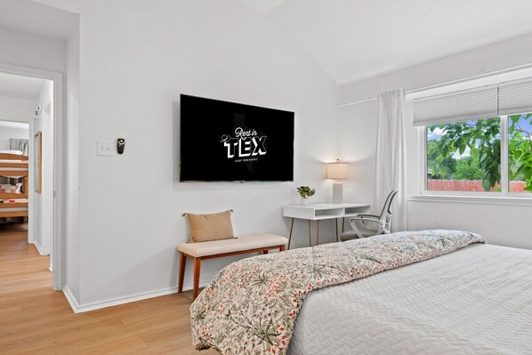 A bedroom with a bed, a desk, a chair, a TV, and a window with a view of greenery.