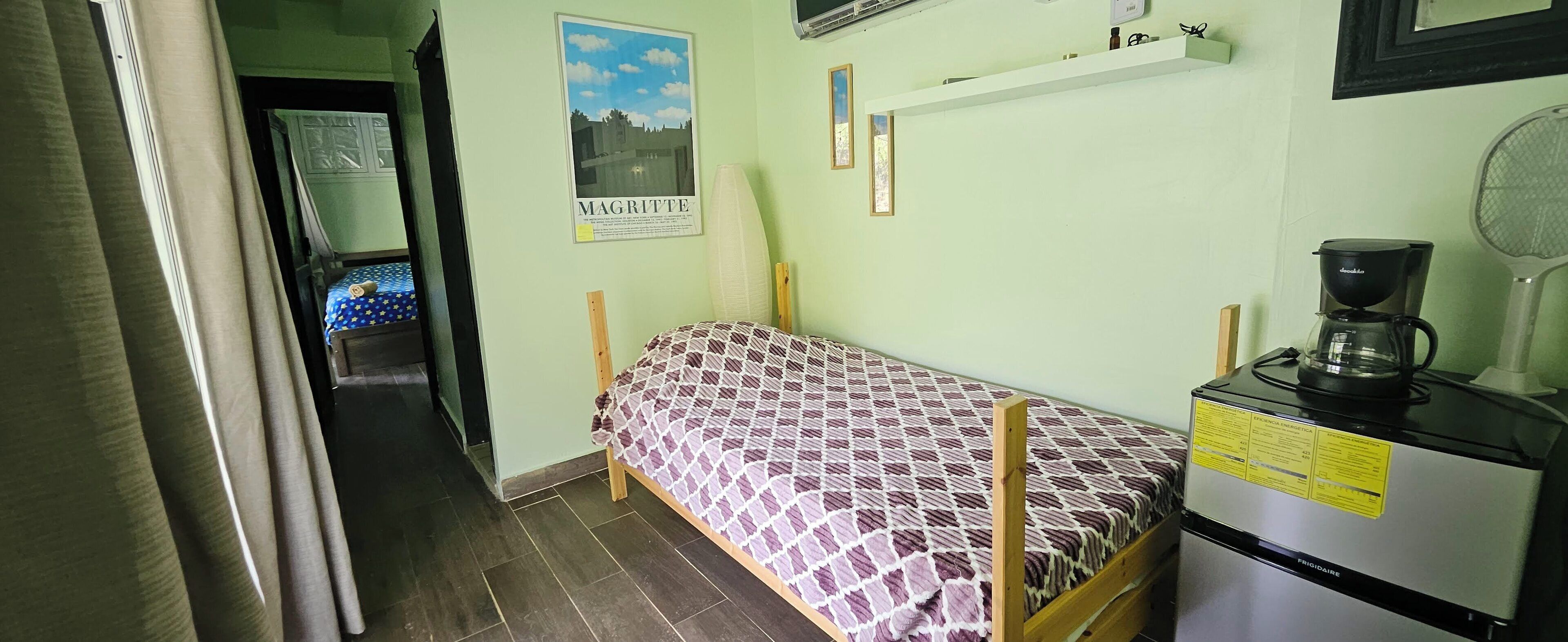 Una habitación pequeña y funcional con una cama individual, una kitchenette con cafetera y un póster en la pared.