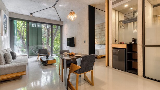 Una sala de estar moderna con sofá, mesa de comedor y sillas, un área de cocina con refrigerador, y ventanas amplias con vista a la vegetación.