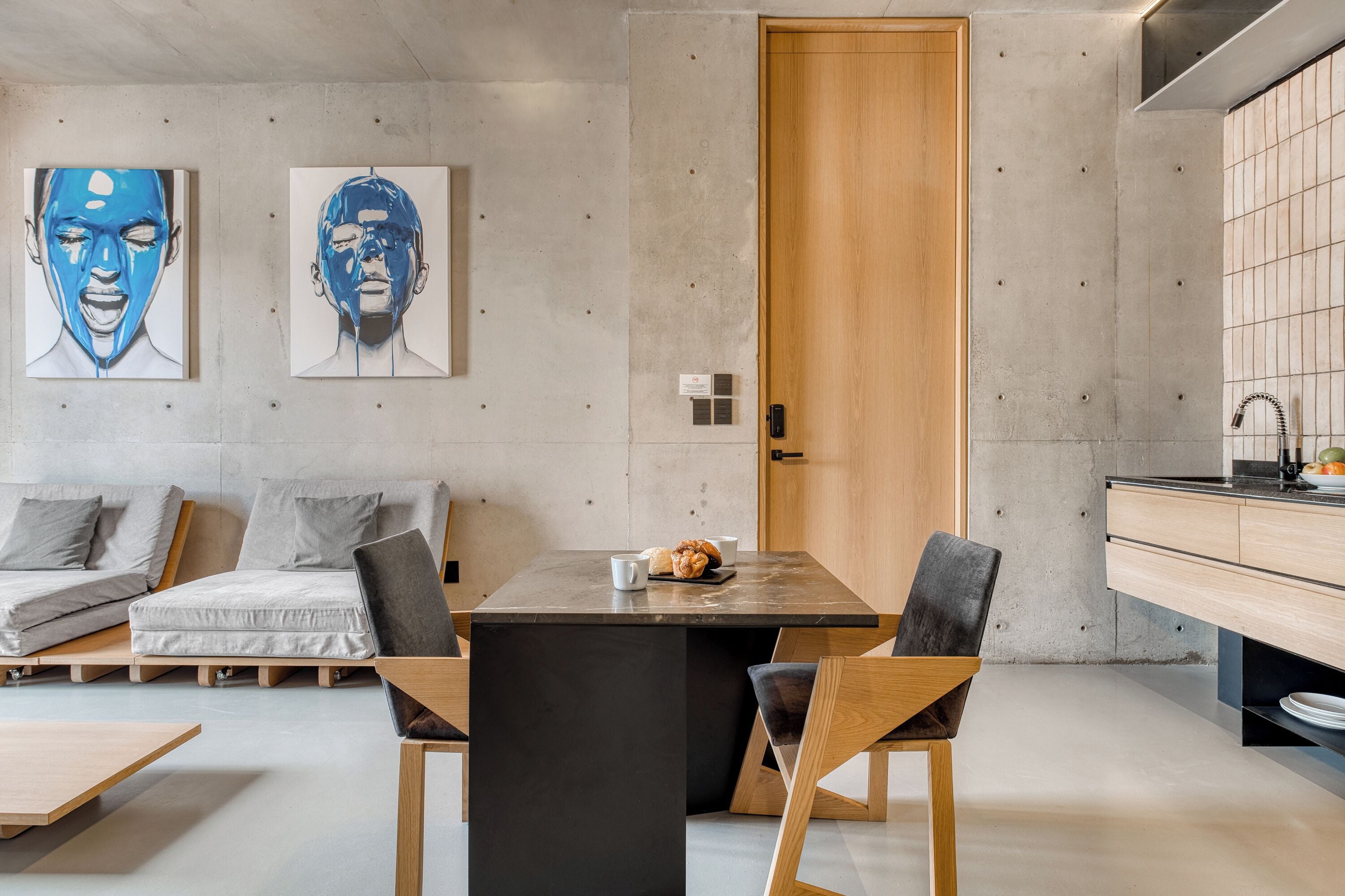 A modern kitchen with a wooden door, a dining table with chairs, and two abstract paintings on the wall.