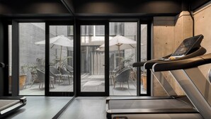 Un gimnasio moderno con una caminadora, una bicicleta estática y un amplio ventanal con vistas a un patio exterior con sombrillas y asientos.