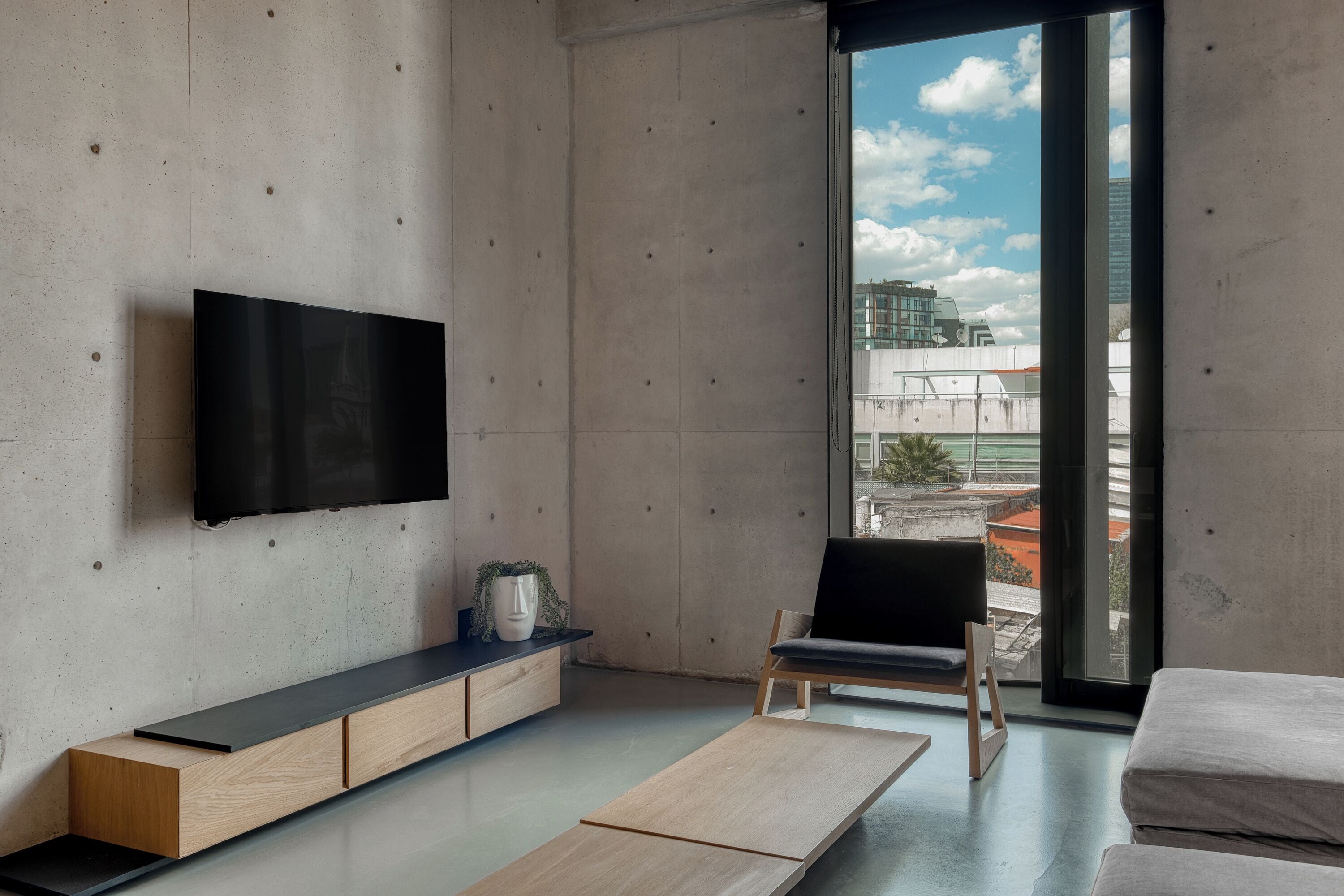 Ein modernes Wohnzimmer mit einem flachbildfernseher an einer Betonwand, einem Holztischchen, einem schwarzen Sessel und einem großen Fenster mit Blick auf Gebäude und einen klaren Himmel.