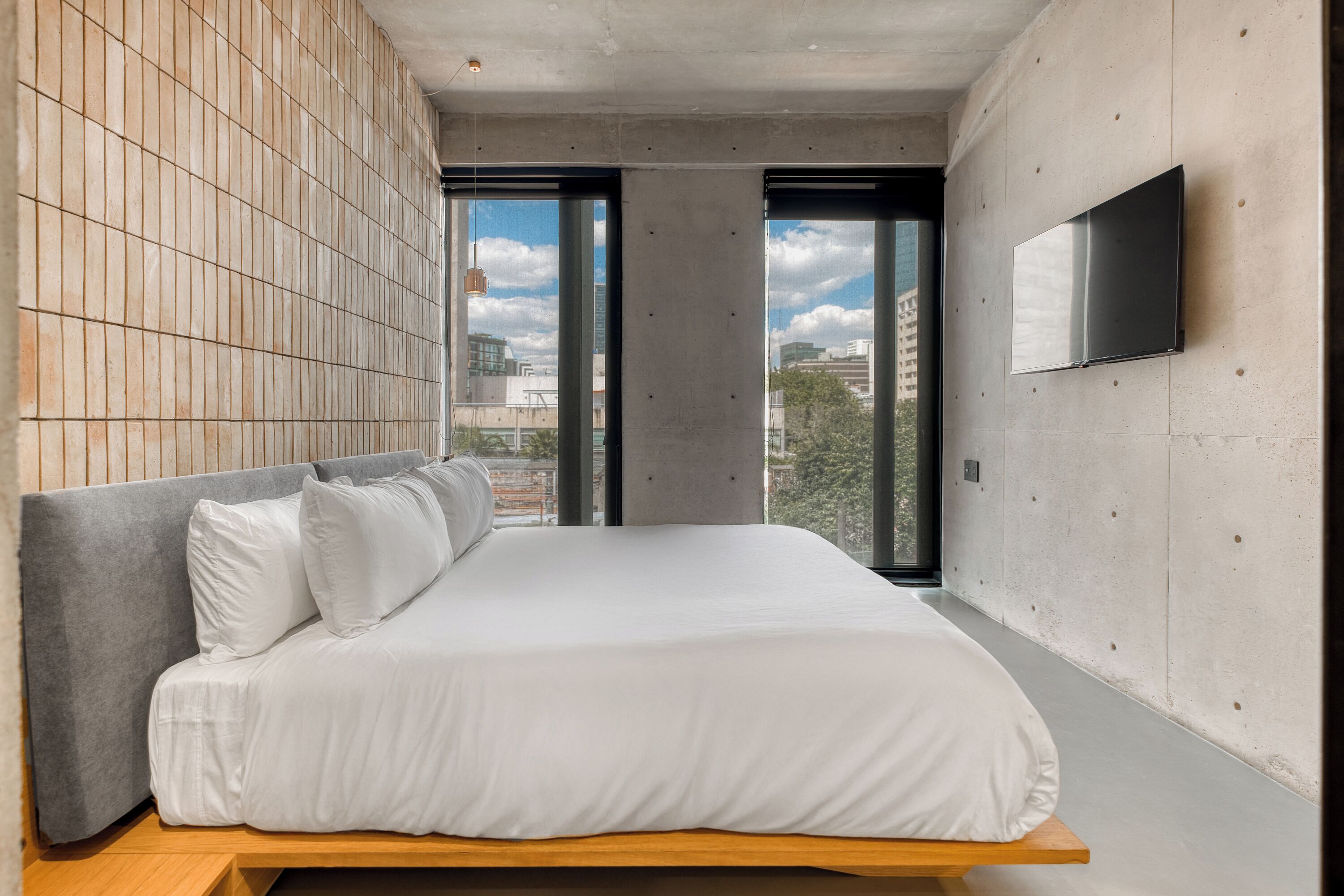 A modern bedroom with a bed, a TV mounted on the wall, and large windows offering a city view.