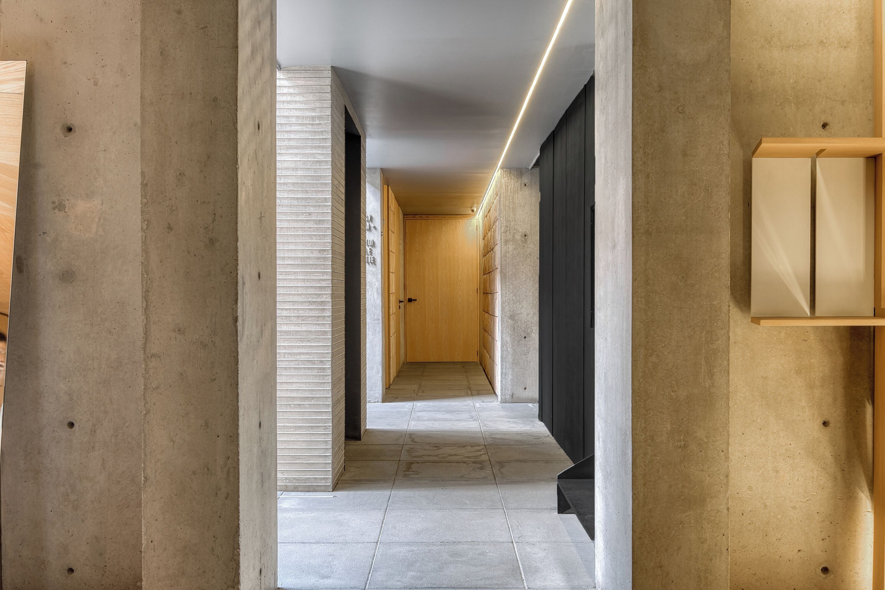 A narrow hallway with a wooden door at the end, tiled floor, and a light strip along the ceiling.