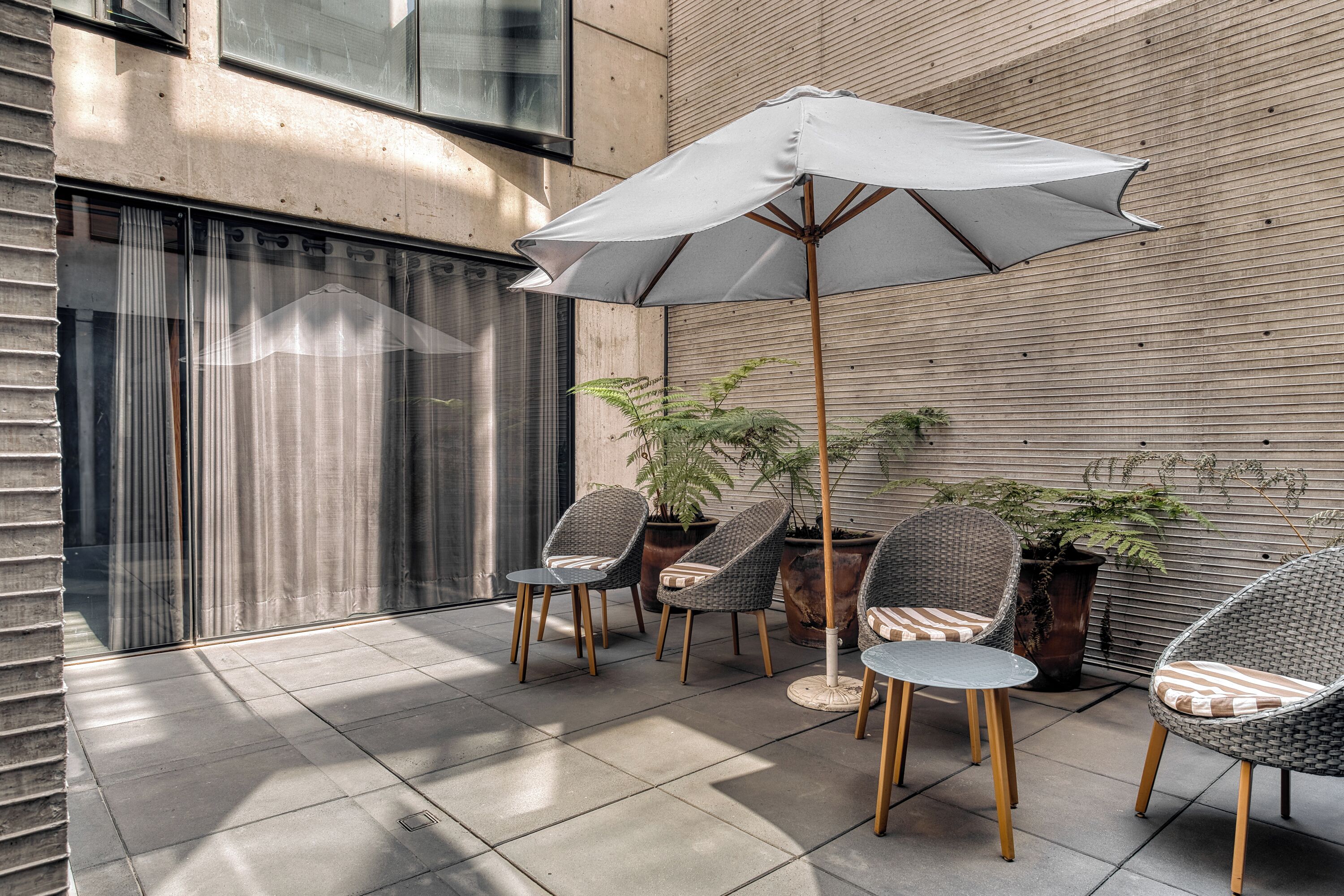 An outdoor seating area with wicker chairs and a table, an umbrella, and potted plants against a building wall.