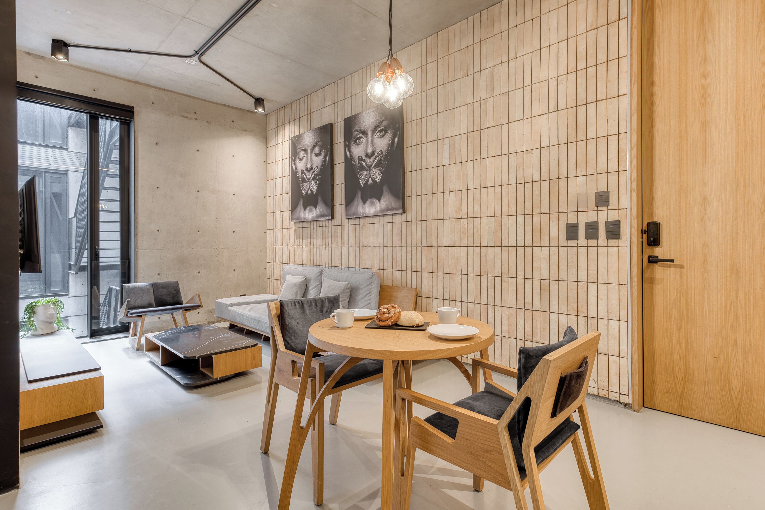 A modern interior with a round wooden table, grey sofa, and wooden chairs. There are two wall-mounted artworks and a wooden door.