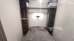 Une petite pièce comprenant un lit, une étagère avec une boîte et une lampe fixée au mur.