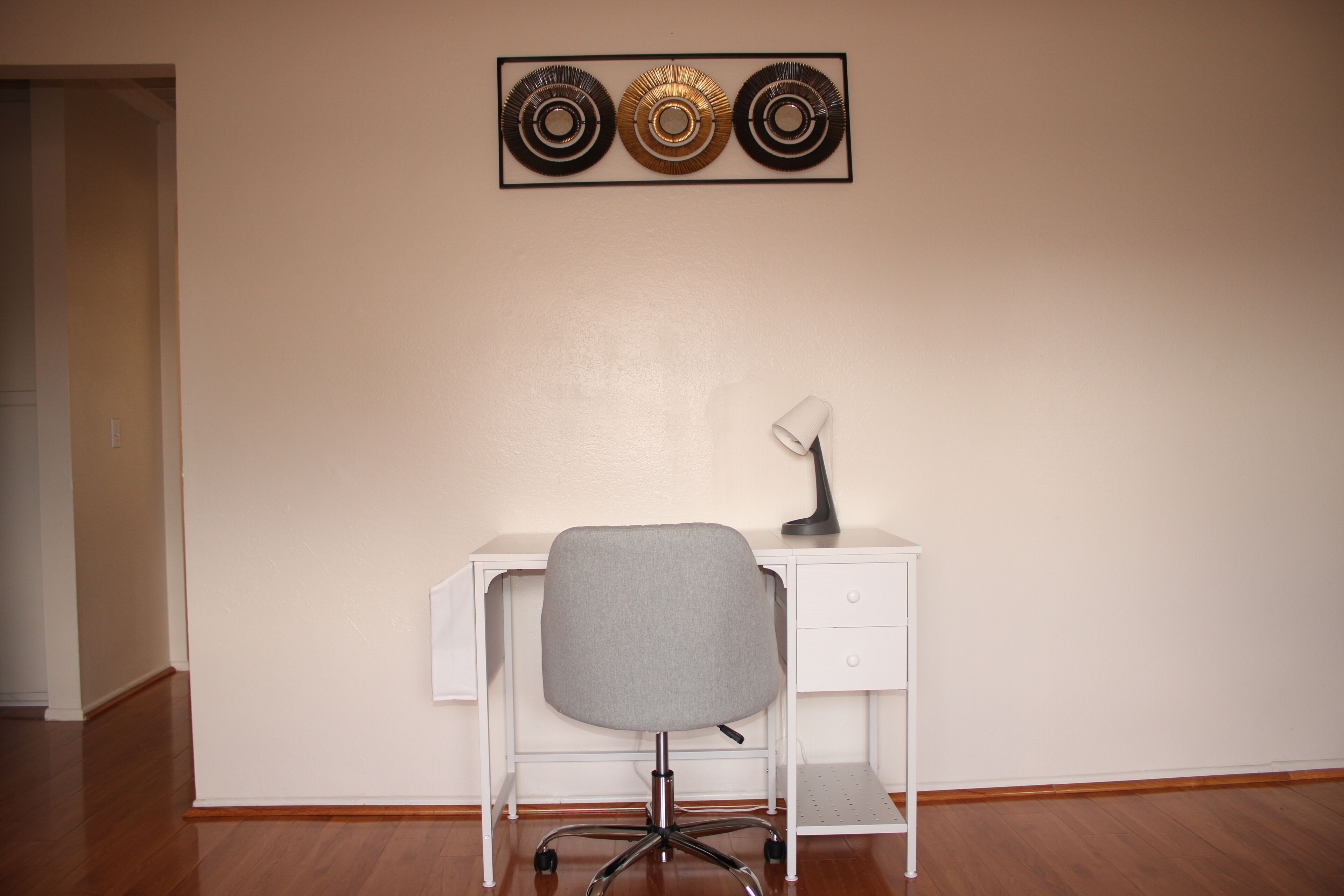 A white desk with a grey office chair, a lamp, and a decorative wall piece.
