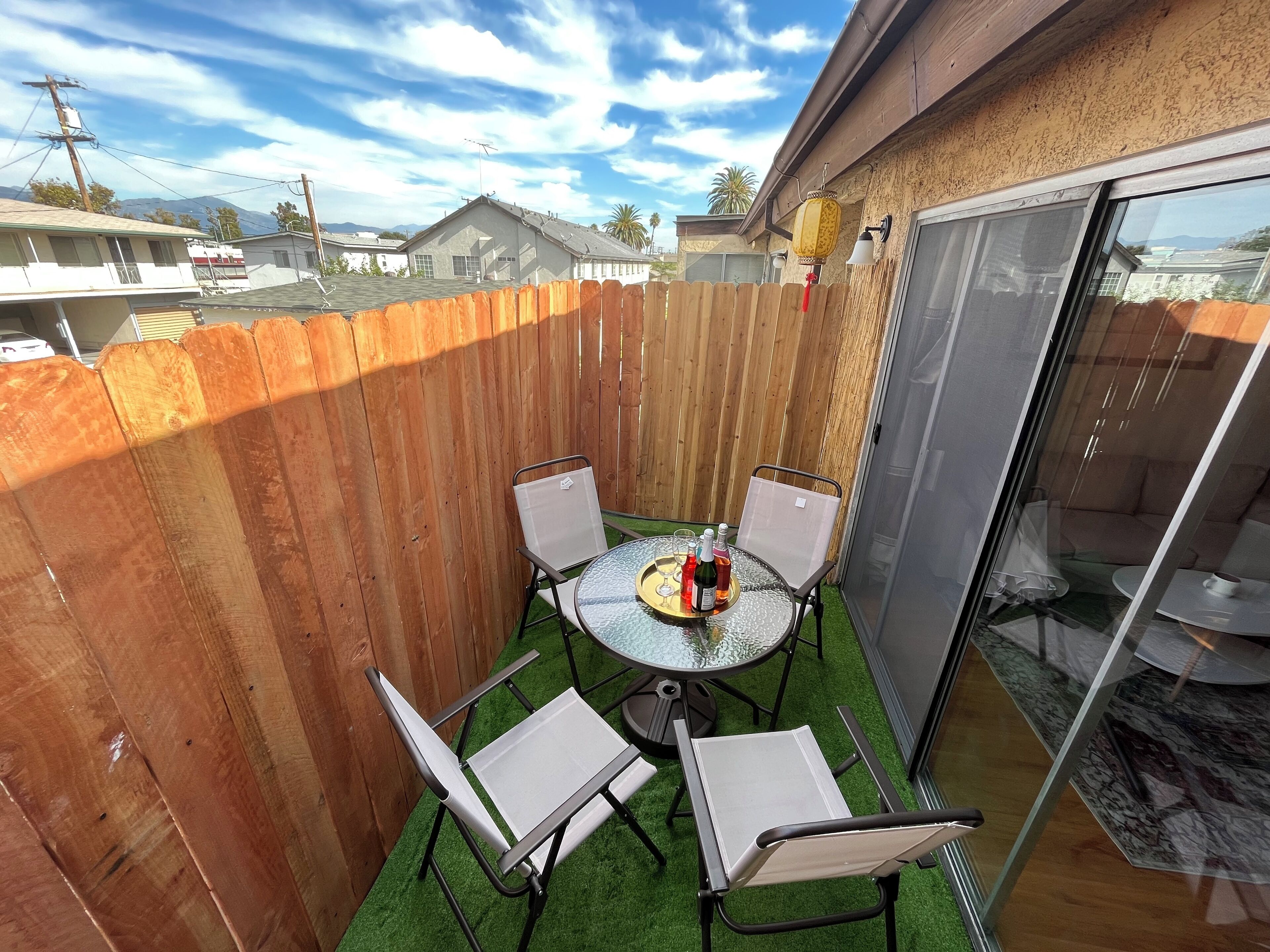 Une terrasse avec une table ronde et quatre chaises, une clôture en bois et une porte vitrée donnant sur une pièce où se trouve un canapé.