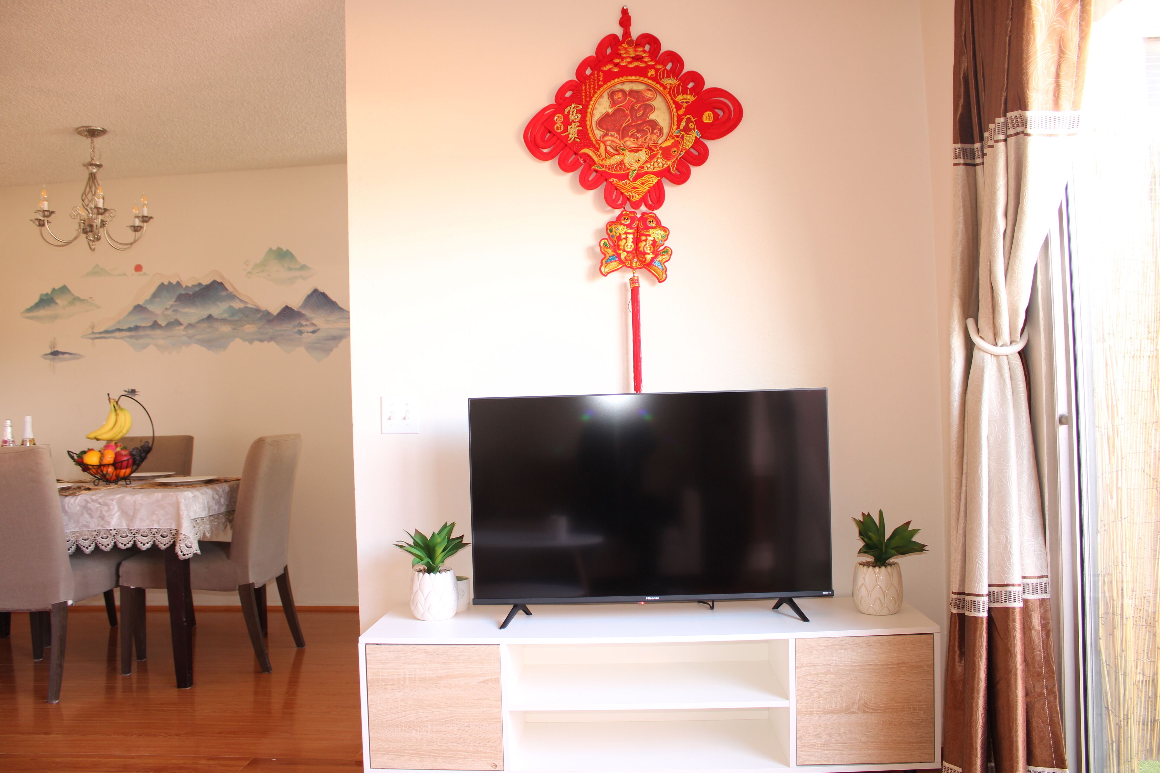 A living room with a flat-screen TV on a white stand, a dining table with chairs, and a decorative wall hanging.