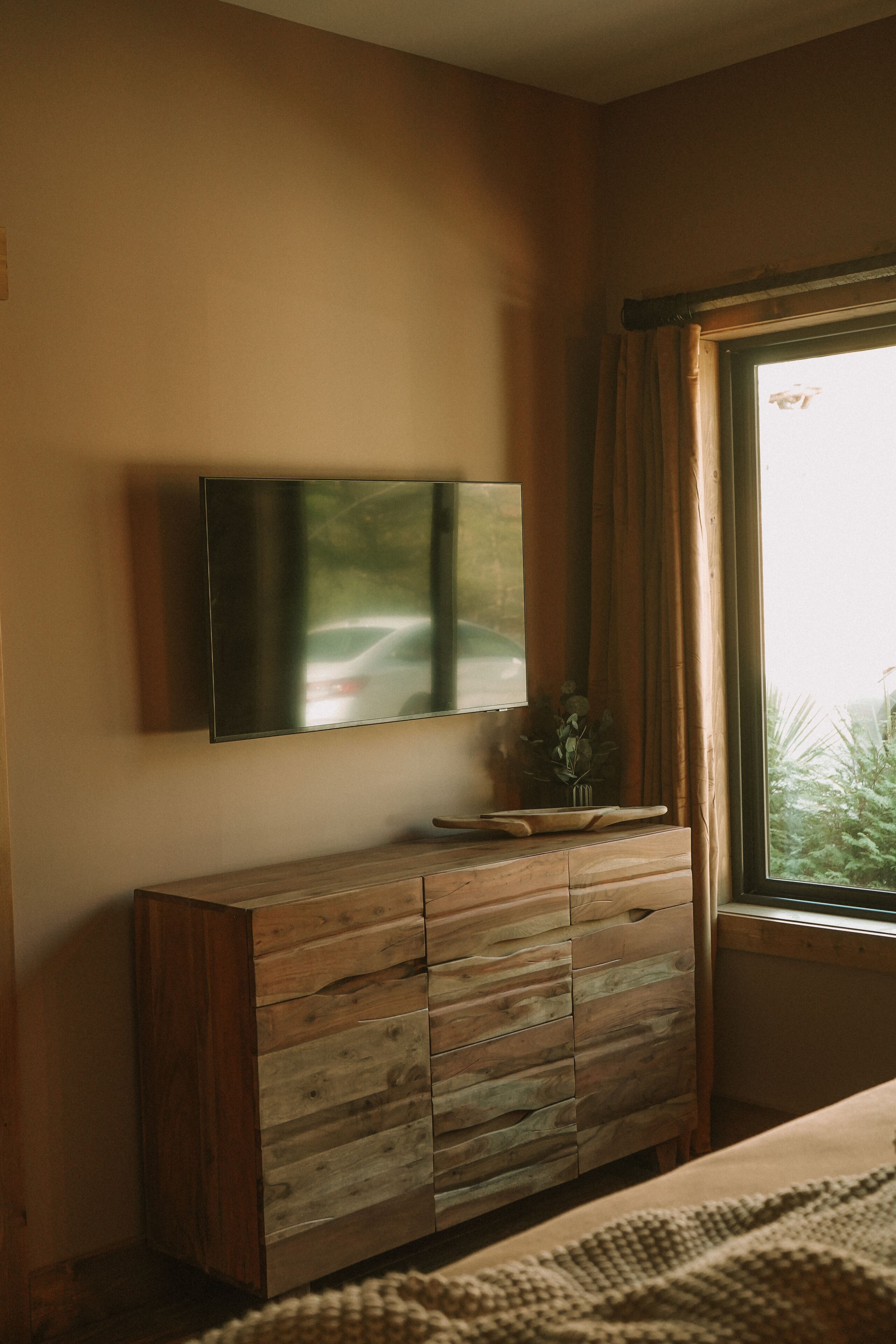 Une chambre à coucher avec une commode en bois, un téléviseur à écran plat fixé au mur et une fenêtre avec des rideaux.