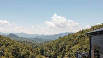 A wooden cabin with a deck overlooking a forested valley and distant mountains.