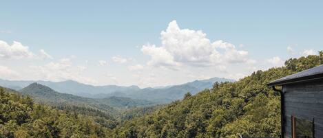 A wooden cabin with a deck overlooking a forested valley and distant mountains.