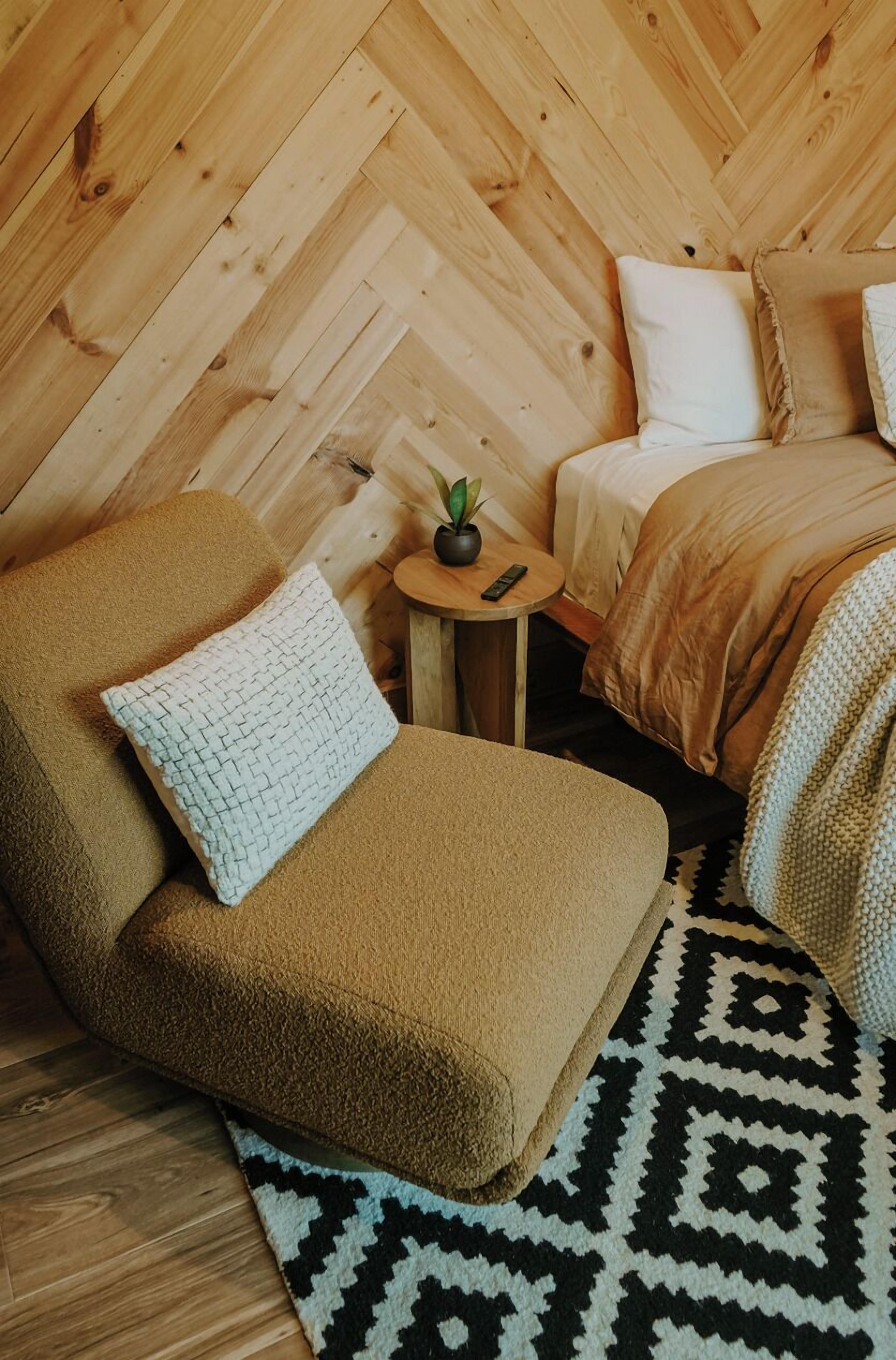 Una habitación acogedora con techo de madera en patrón espiga, un sofá marrón con un cojín texturizado, una mesita redonda con una planta y una alfombra estampada.
