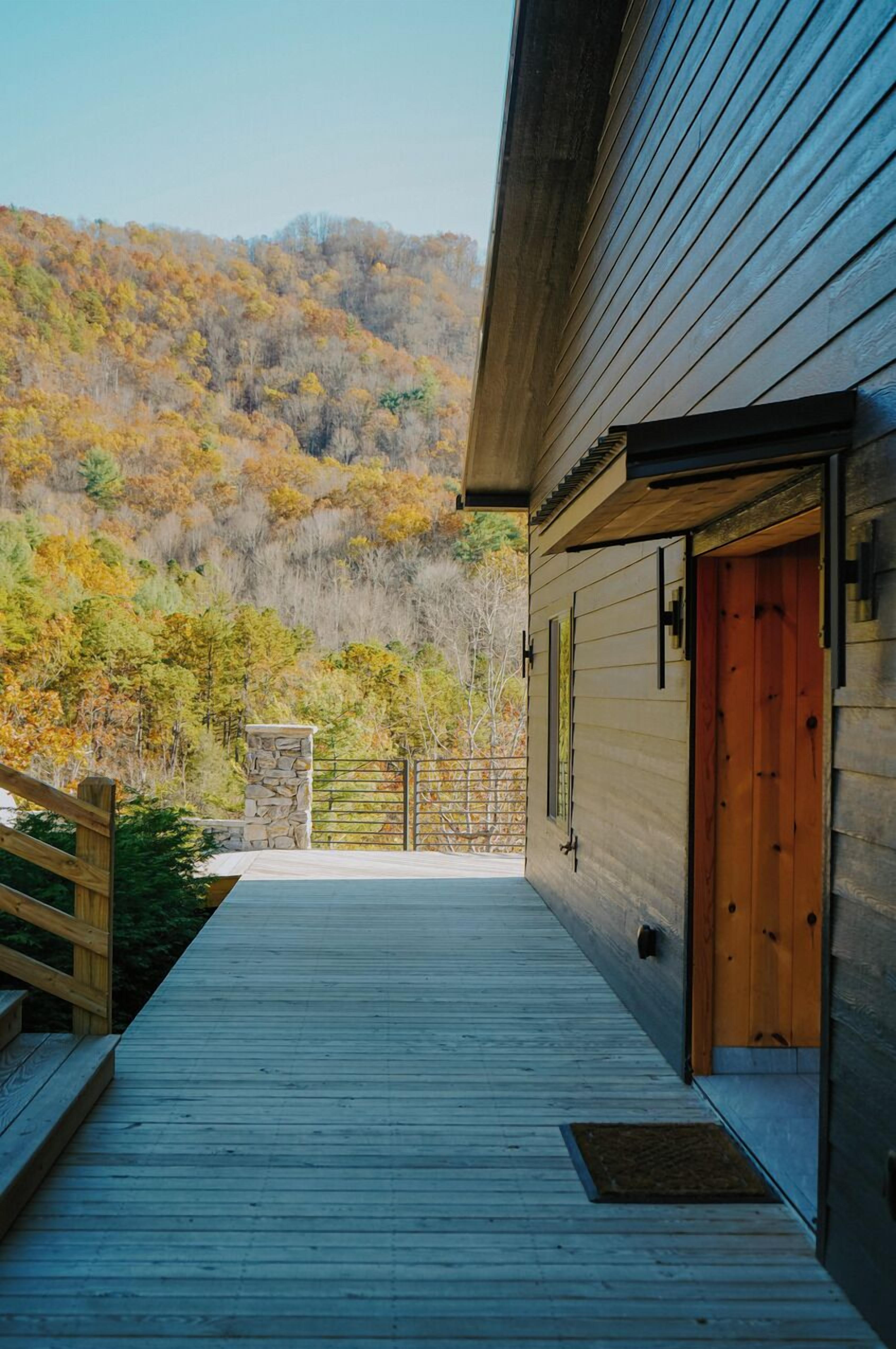 Une terrasse en bois mène à un bâtiment doté d’un pilier en pierre et d’une porte en bois.