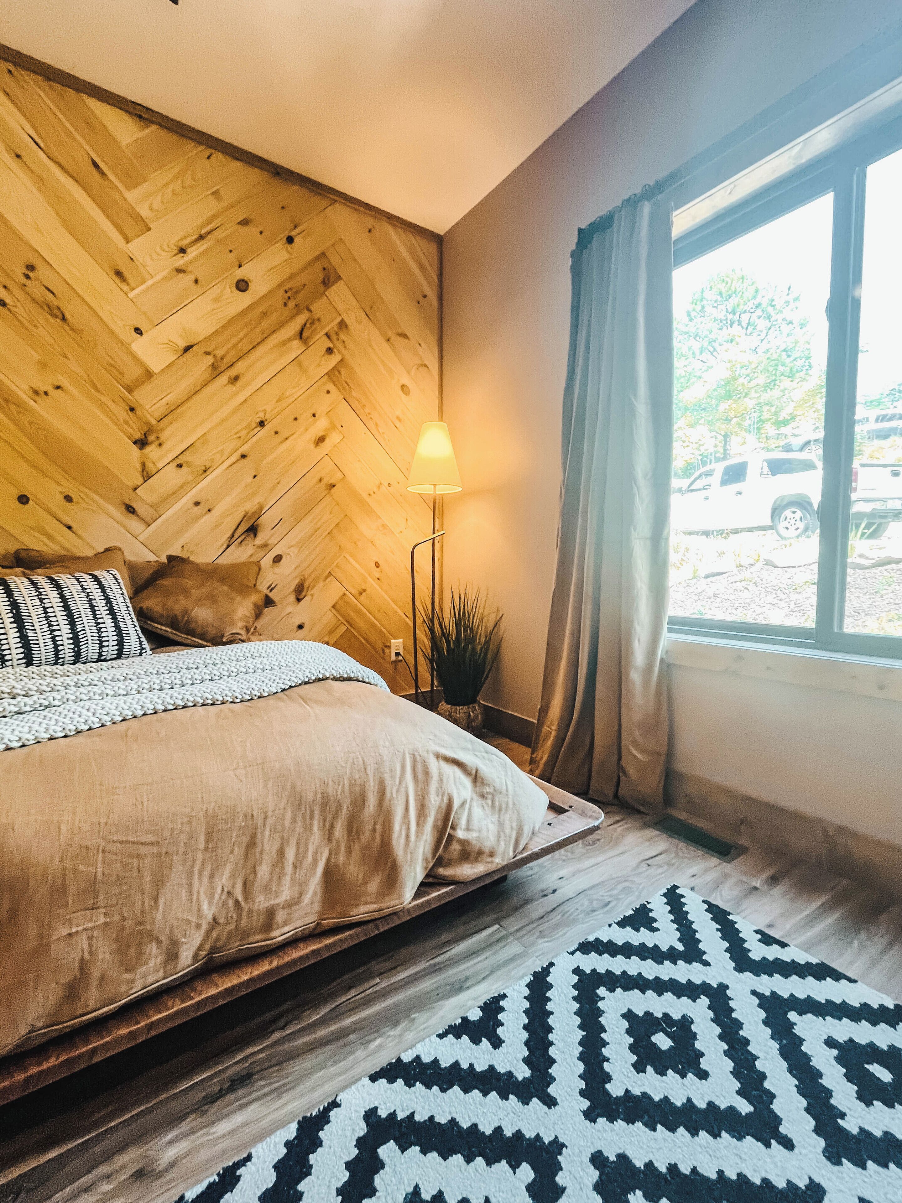 Een slaapkamer met een wand van visgraat-hout, een bed met een beddekleed met een patroon, een bedlamp en uitzicht op geparkeerd vervoer buiten.