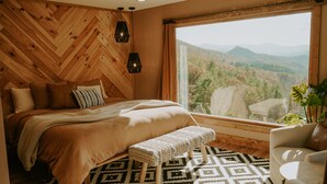 Een slaapkamer met een groot raam, houten lambrisering, een bed met een geborduurd plaid, een bankje en uitzicht op een bebost berglandschap.