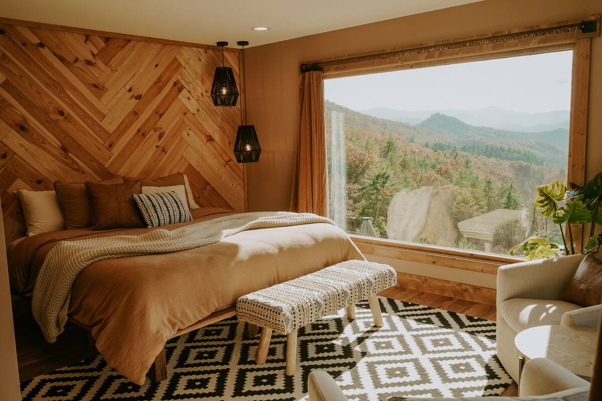 Un dormitorio con un ventanal grande, paneles de madera en la pared, una cama con manta estampada, un banco y vistas a una cadena montañosa boscosa.